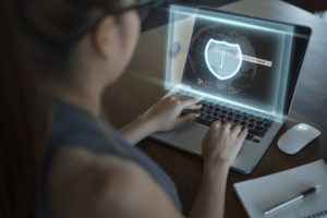 A dark-haired woman wearing glasses works on a laptop that displays a glowing light blue shield on the screen to symbolize cybersecurity.