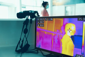 A thermal camera with a display screen in the foreground is used to gauge the body temperature of a woman wearing a medical mask in the background.