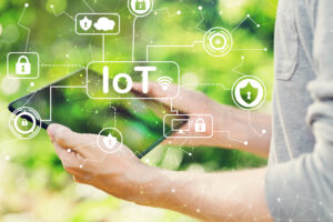 A person holds a tablet against a green, leafy, blurred background with the initials IoT superimposed over the scene, along with symbols like a shield to represent IoT security.