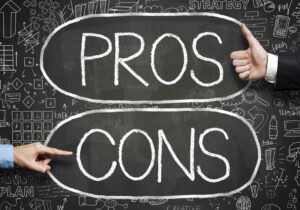 A blackboard has PROS and CONS written on it in white chalk, with a hand pointing to CONS and another hand giving a thumbs up next to PROS.