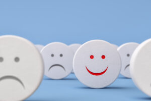 A smiley face stands among a crowd of frowny faces against a pale blue background.