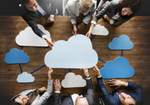 A group of people in business attire hold a paper cutout of a cloud over a conference table, symbolizing collaboration through cloud computing.