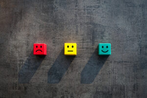 A red block with a frowning face on it, a yellow block with a neutral face, and a green block with a smiling face against a gray background, symbolizing customer experience.
