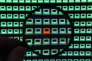 Close up of a person holding a magnifying glass over a screen filled with tiny green computer icons and one red computer at the center of the screen.