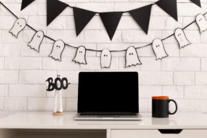 Picture of a desk with a laptop and black mug. On the white brick wall above the desks, there's a garland of ghosts, and there's a ghost figurine on the desk holding the word "Boo."