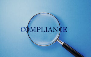 A magnifying glass placed over the word "compliance" against a blue background.