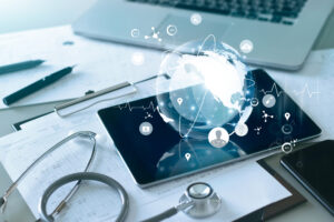 A stethoscope, tablet and laptop on a desk with a glowing globe icon floating over the tablet, symbolizing healthcare communications. 