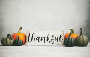 A sign that says "thankful" in cursive flanked by orange and green pumpkins against a pale gray background.