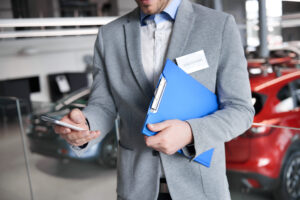 Picture of a car salesperson holding a folder in one hand and a cell phone in the other, with cars in the background behind them.
