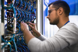Side view portrait of young man connecting cables in server cabinet while working with supercomputer in data center.