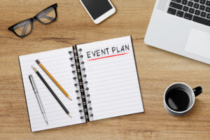 An open spiral notebook that says "EVENT PLAN" across the top of one page lies on a desk beside a pair of glasses, a smartphone, a cup of coffee, and a laptop.