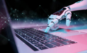 A robot hand extends its index finger to press the space bar on a laptop keyboard, representing artificial intelligence.
