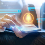 Tech Talks: Contact center voice biometrics can save $400K annually Close up of a person using a smartphone with a glowing microphone symbol hovering over the screen.