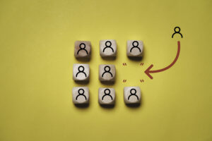 Against a yellow background, blocks with line drawings symbolizing people sit in three rows (one row of three, one row of two, and a final row of three blocks). An arrow points from a separate line drawing symbolizing a person to the empty space in the middle row of two blocks, representing hiring or recruiting.