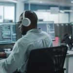 Tech Talks: How a Managed Services Partner Helps IT Leaders Strengthen Security, IT Support, and Compliance Office: Professional Black IT Programmer Uses Headphones while Working on Desktop Computer. Male Website Developer, Software Engineer Developing App, Video Game. Listening to Podcast, Music.